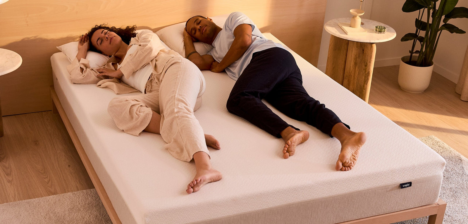 Couple allongé en position fœtale sur un matelas mousse Wopilo posé sur un lit en bois clair, dans une chambre lumineuse avec tables de chevet, lampes et plante verte.