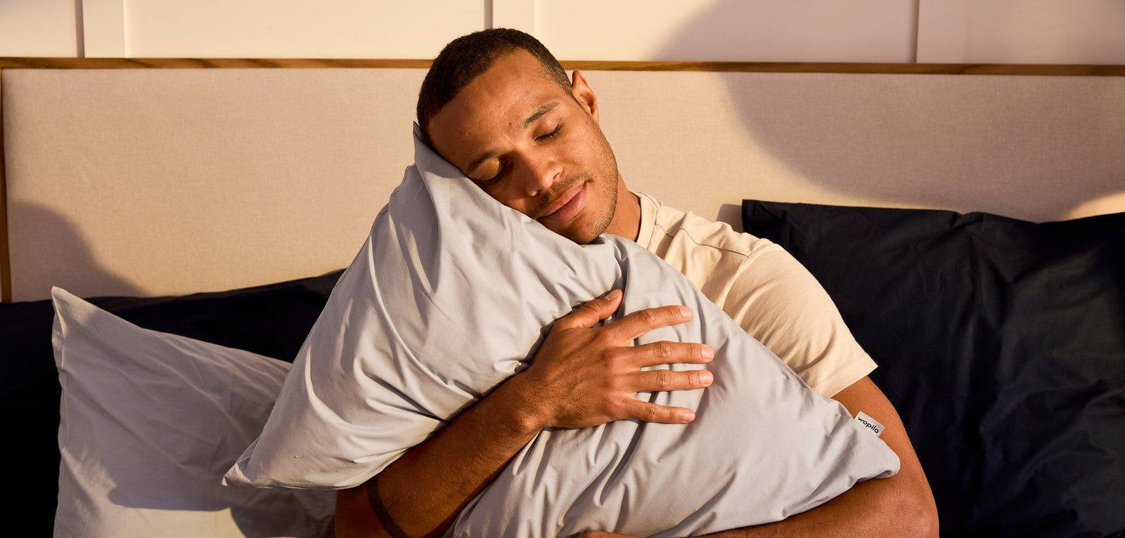 Un homme assoupi sereinement en serrant un oreiller bleu clair contre lui, la tête posée dessus, dans une chambre éclairée par une lumière chaude et apaisante.