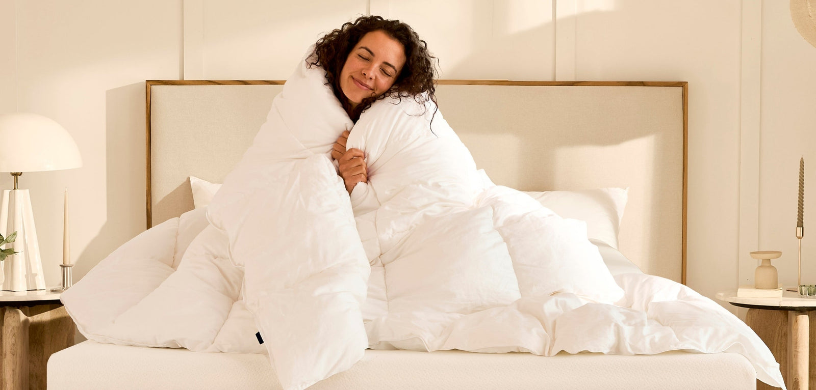 Une femme aux cheveux bouclés, assise sur son lit, s'enroule avec bonheur dans une grande couette blanche épaisse, illustrant le confort et le bien-être .