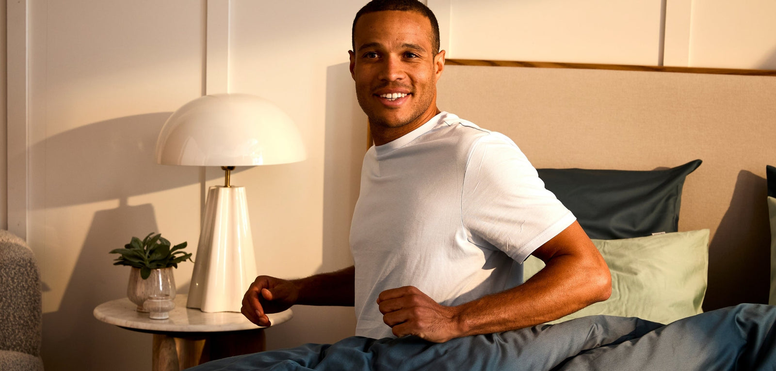 Un homme souriant venant de ce réveiller, assis dans au bord de son lit avec une couette bleue, dans une chambre lumineuse 