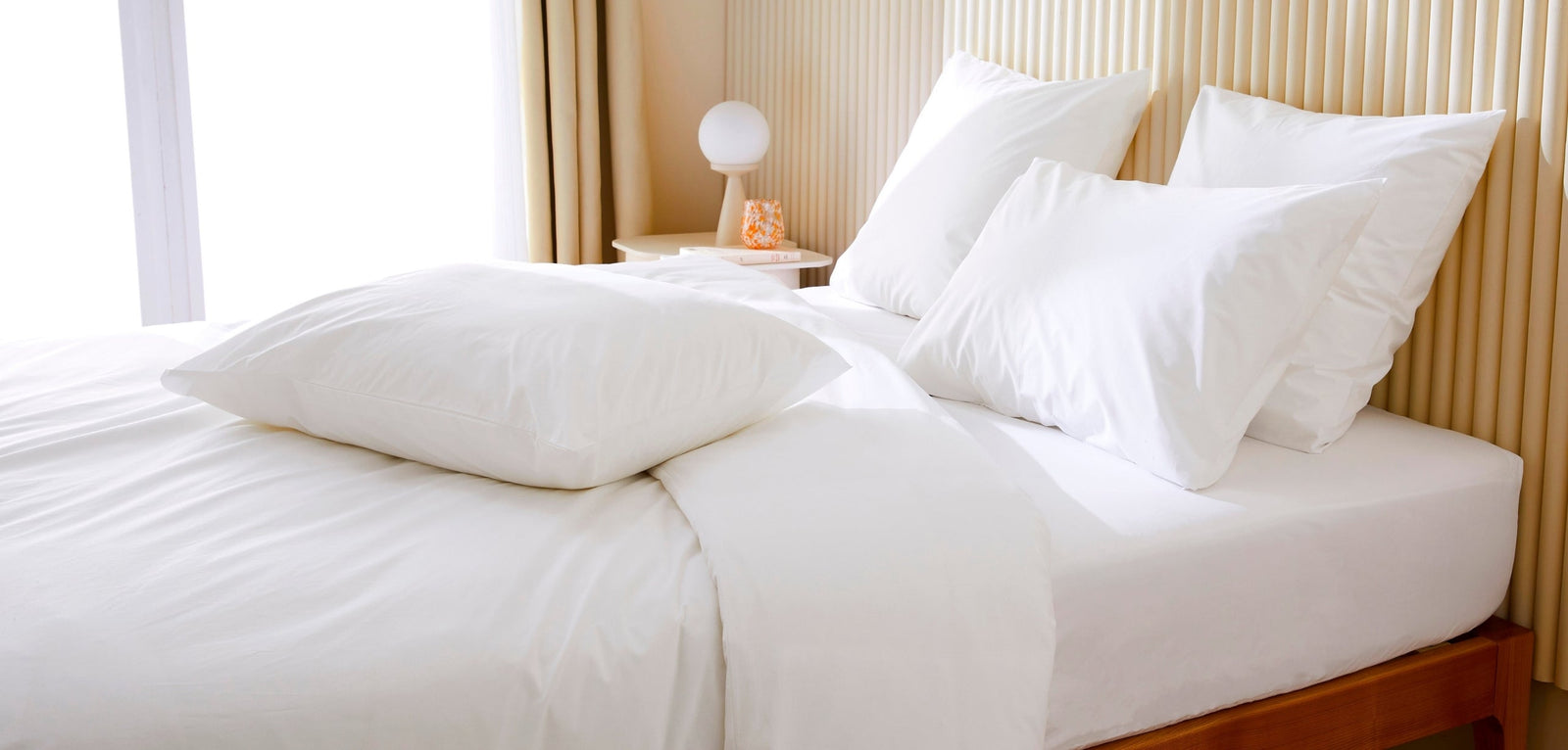 Une parure de lit d'un blanc éclatant comprenant des oreillers, une couette et un drap-housse, installée dans une chambre lumineuse avec une tête de lit en lattes de bois clair, illustrant la fraîcheur et la propreté.