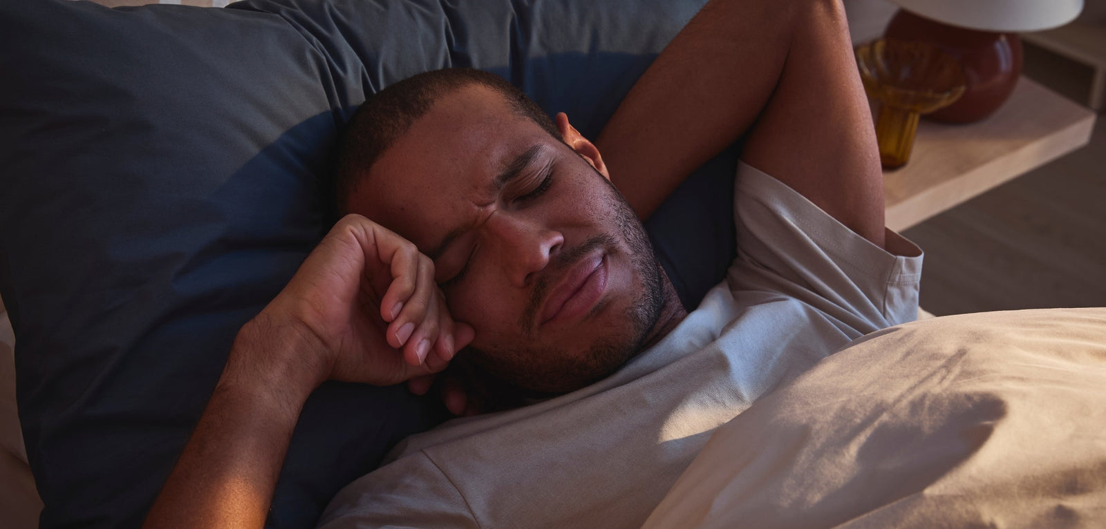 Homme allongé dans son lit, les yeux fermés et le visage légèrement crispé, la tête posée sur un oreiller sombre, partiellement couvert par une couette claire, dans une chambre faiblement éclairée.