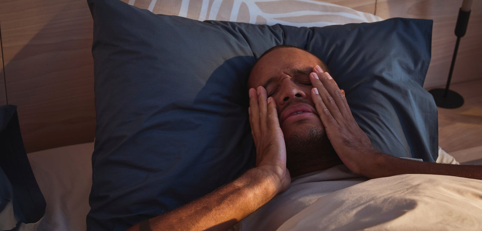 Homme allongé dans son lit, les yeux fermés, les mains sur le visage, exprimant de la fatigue au réveil. Il repose sur un oreiller bleu dans une chambre à la lumière douce du matin.