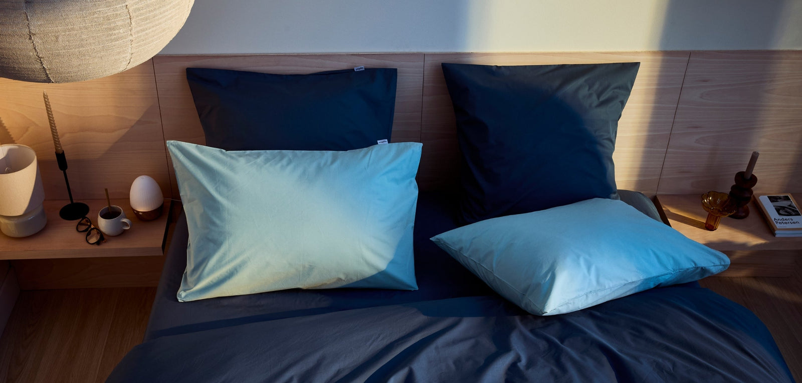 Lit avec draps et taies d’oreiller bleu foncé et bleu clair, disposé dans une chambre chaleureuse avec tête de lit en bois et tables de chevet décorées d’objets et lampes.