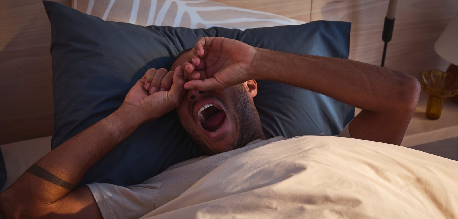 Un homme dans son lit qui baille aux éclats en se frottant les yeux, illustrant la fatigue, sur un oreiller bleu foncé.