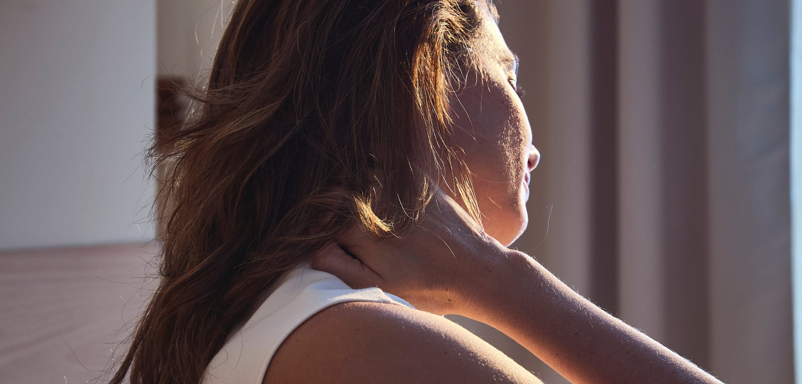 Gros plan de profil d'une femme se massant délicatement la nuque avec la main, illustrant une tension ou une douleur aux cervicales dans une lumière matinale douce.