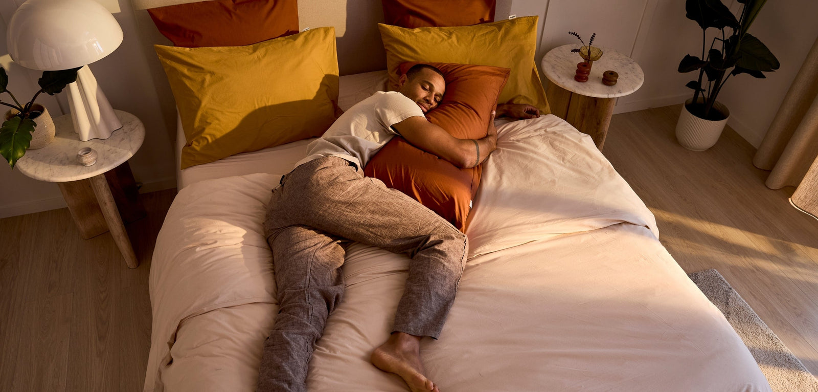 Vue de haut d'un homme endormi sur le côté, enlaçant un grand oreiller terre cuite sur un lit aux tons ocre et beige, baigné par une lumière latérale.