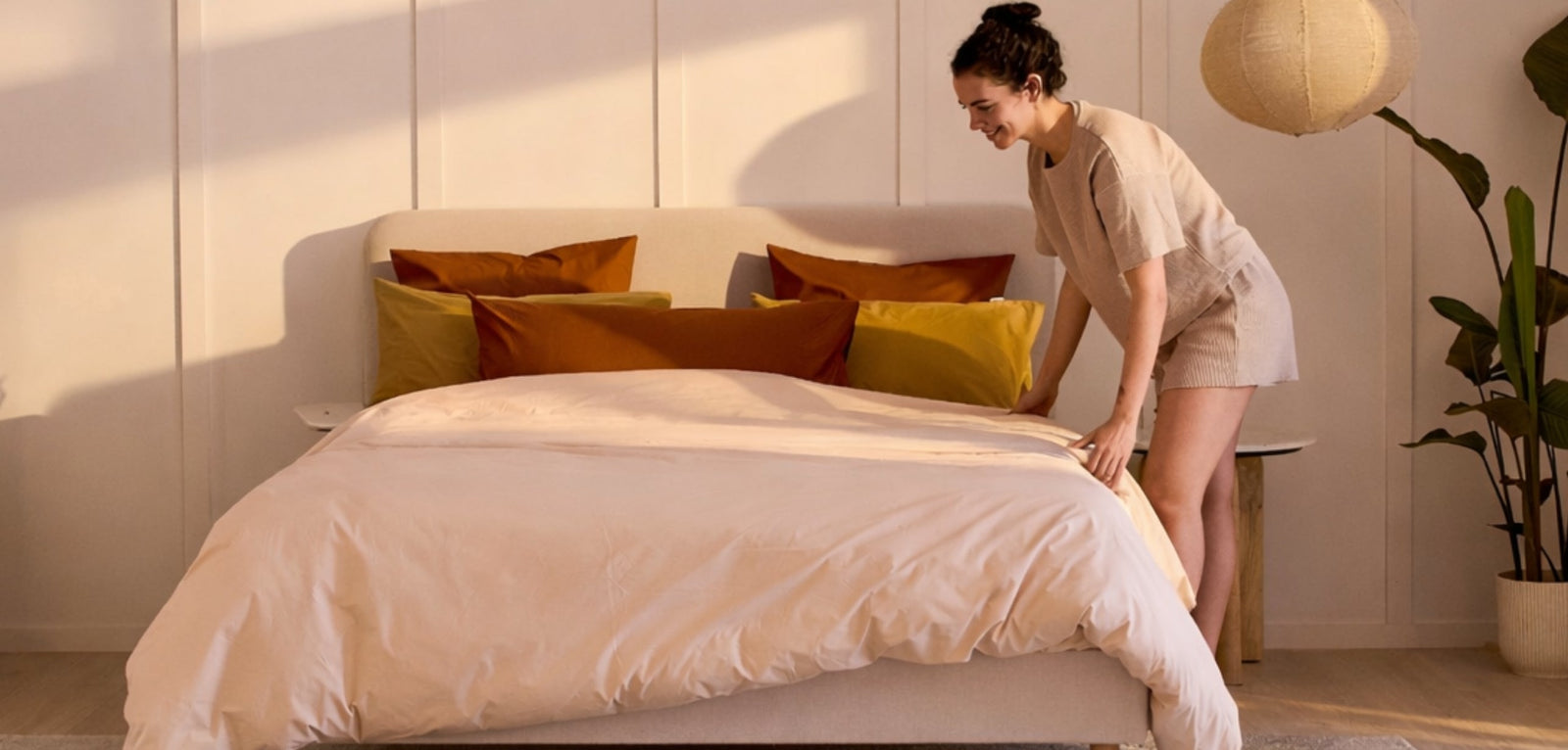 Une femme souriante en train de border une couette beige sur un grand lit, dans une chambre baignée par la lumière chaude du couchant, avec des oreillers aux tons terracotta et moutarde.