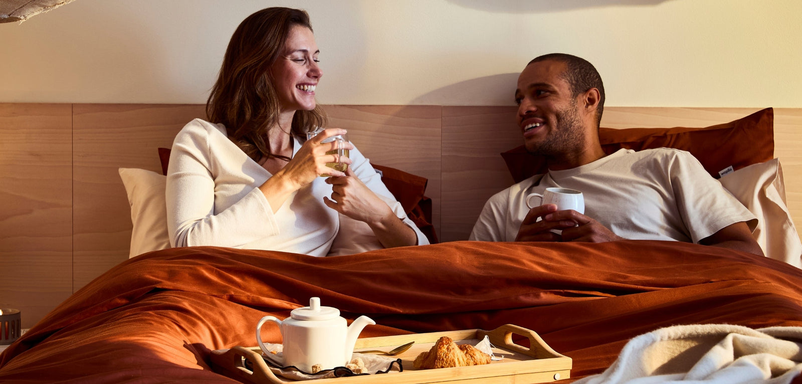 Un homme et une femme assis dans leur lit, discutant et souriant en prenant un petit-déjeuner avec un plateau de thé et croissant, sous une lumière matinale douce.