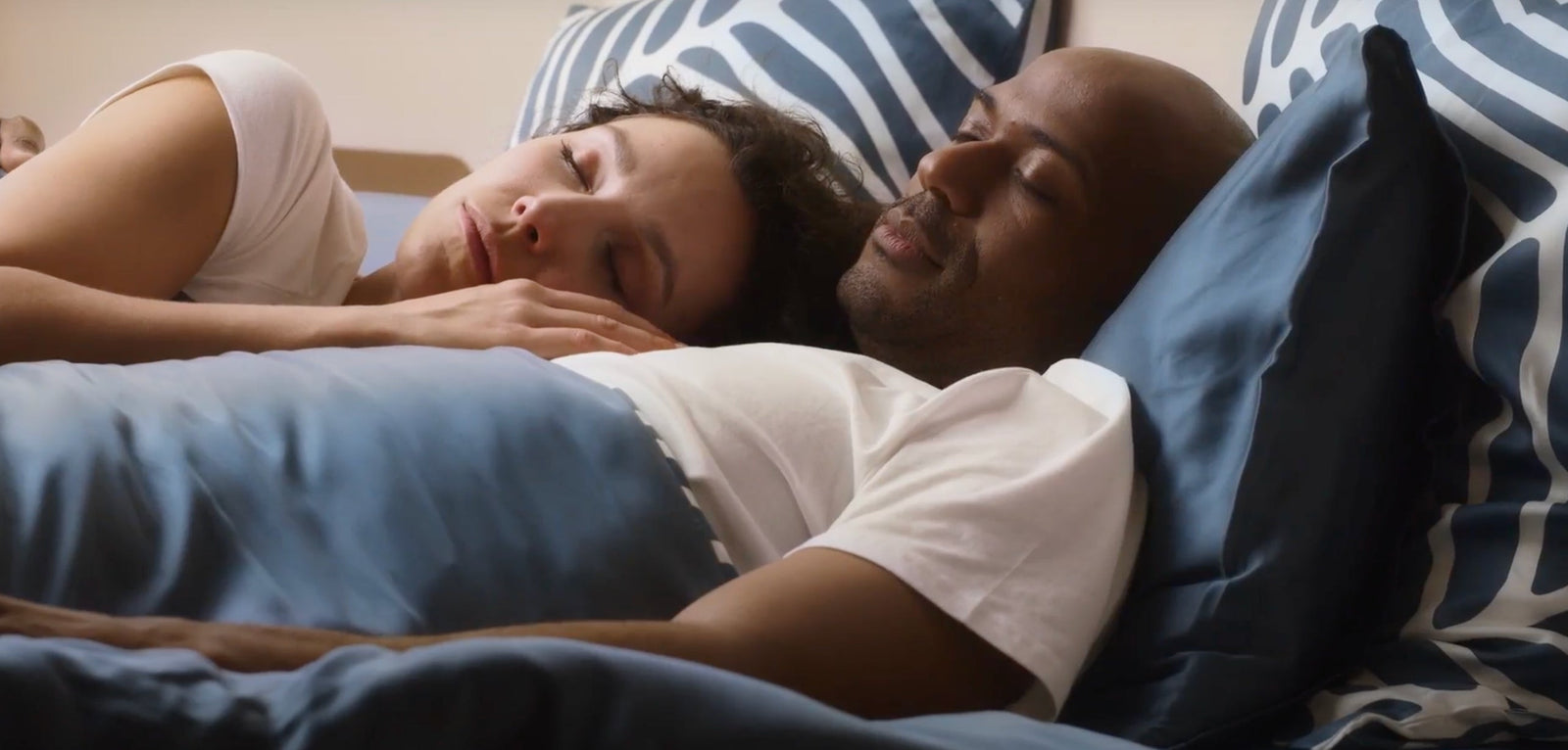 Un couple endormi paisiblement côte à côte sous une couette bleue, la tête reposant sur des oreillers aux motifs graphiques bleus et blancs.