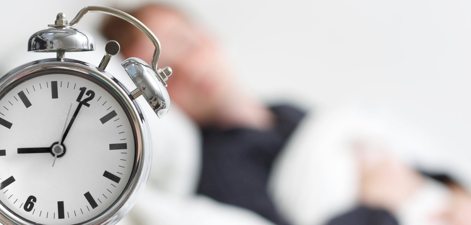 Au premier plan on vois un réveil  sur la gauche, et en fond flouté on peut voir une personne qui dort avec une couette sur elle. 