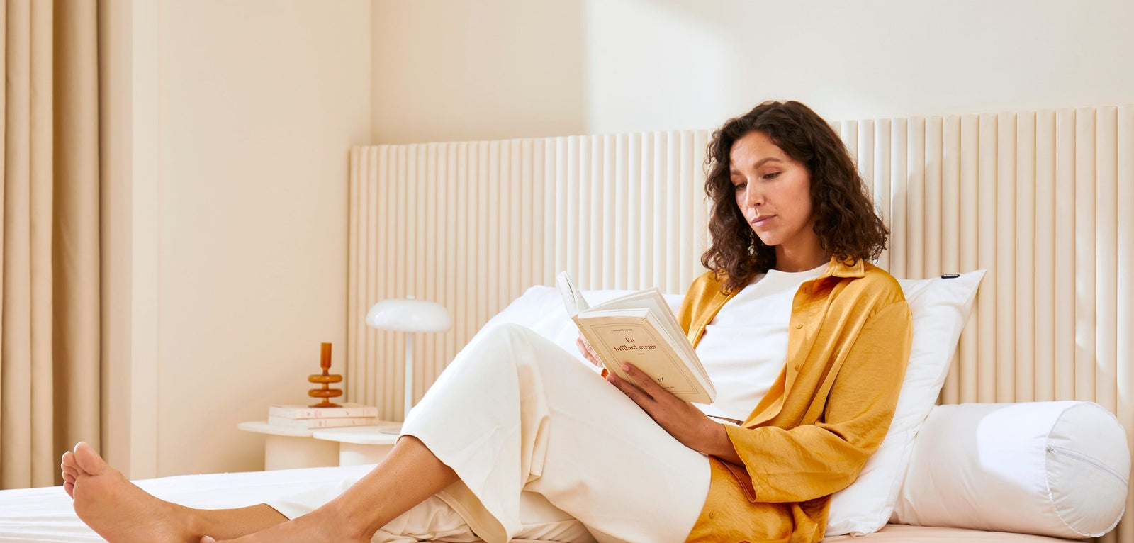Femme assise sur un lit, adossée à des oreillers blancs, lisant un livre dans une chambre lumineuse aux tons beige et bois clair, avec une table de chevet et une lampe en arrière-plan.