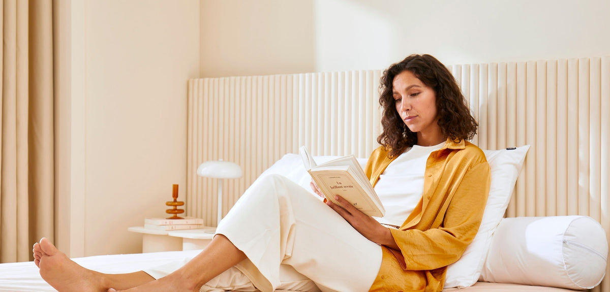 Femme assise sur un lit, adossée à des oreillers blancs, lisant un livre dans une chambre lumineuse aux tons beige et bois clair, avec une table de chevet et une lampe en arrière-plan.