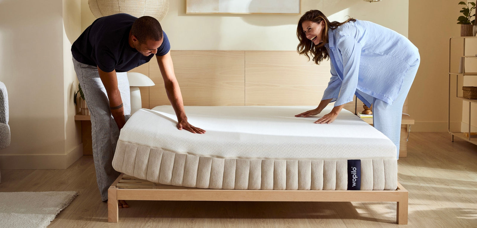Un homme et une femme souriants installent un matelas blanc sur un sommier en bois dans une chambre lumineuse au style épuré. Le matelas, posé sur un cadre de lit en bois clair.