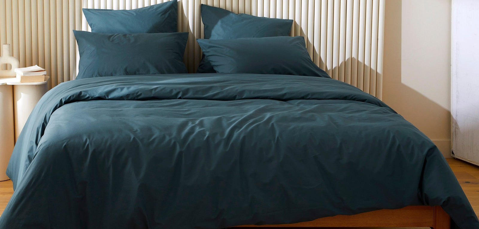 : Une parure de lit complète de couleur bleu canard, comprenant une housse de couette et des oreillers parfaitement lisses et sans plis, sur un lit avec une tête de lit en lattes de bois clair.