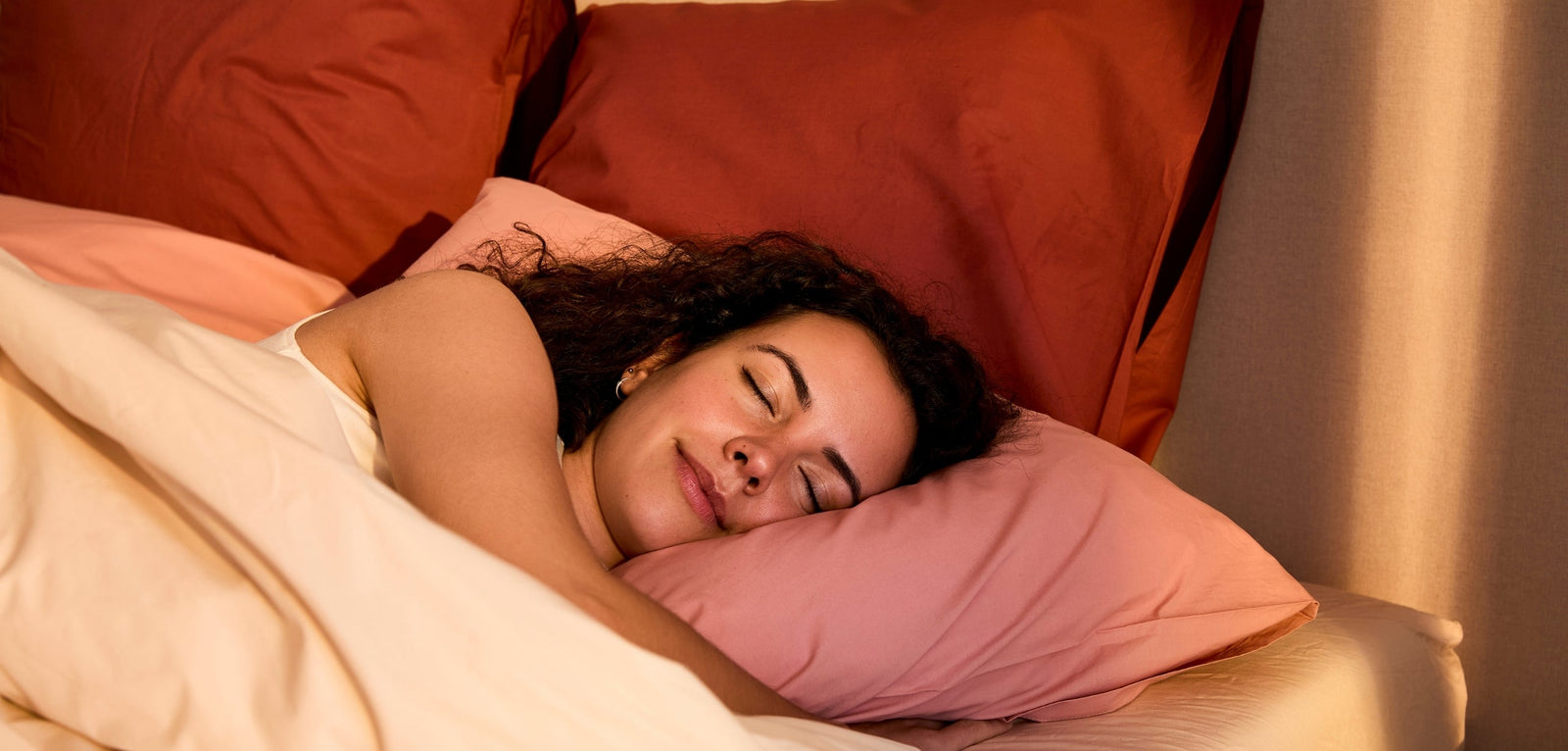 Une femme aux cheveux bouclés endormie paisiblement sur le côté, la tête posée sur un oreiller rose, dans une ambiance chaleureuse avec des oreillers terracotta en arrière-plan.