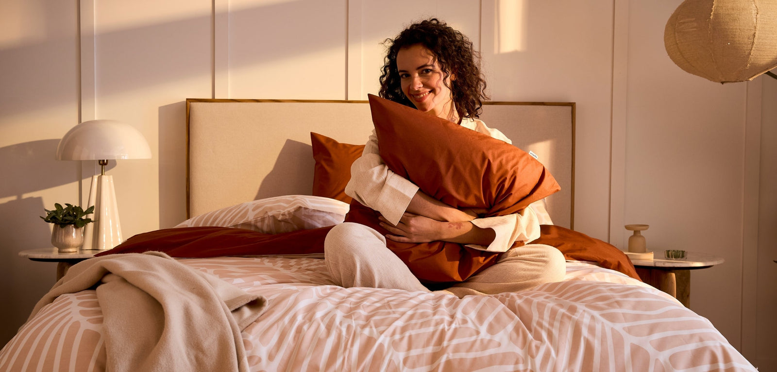 Une femme souriante aux cheveux bouclés, assise sur un lit et serrant un oreiller de couleur terracotta contre elle, dans une chambre baignée par la lumière chaude du soleil couchant.