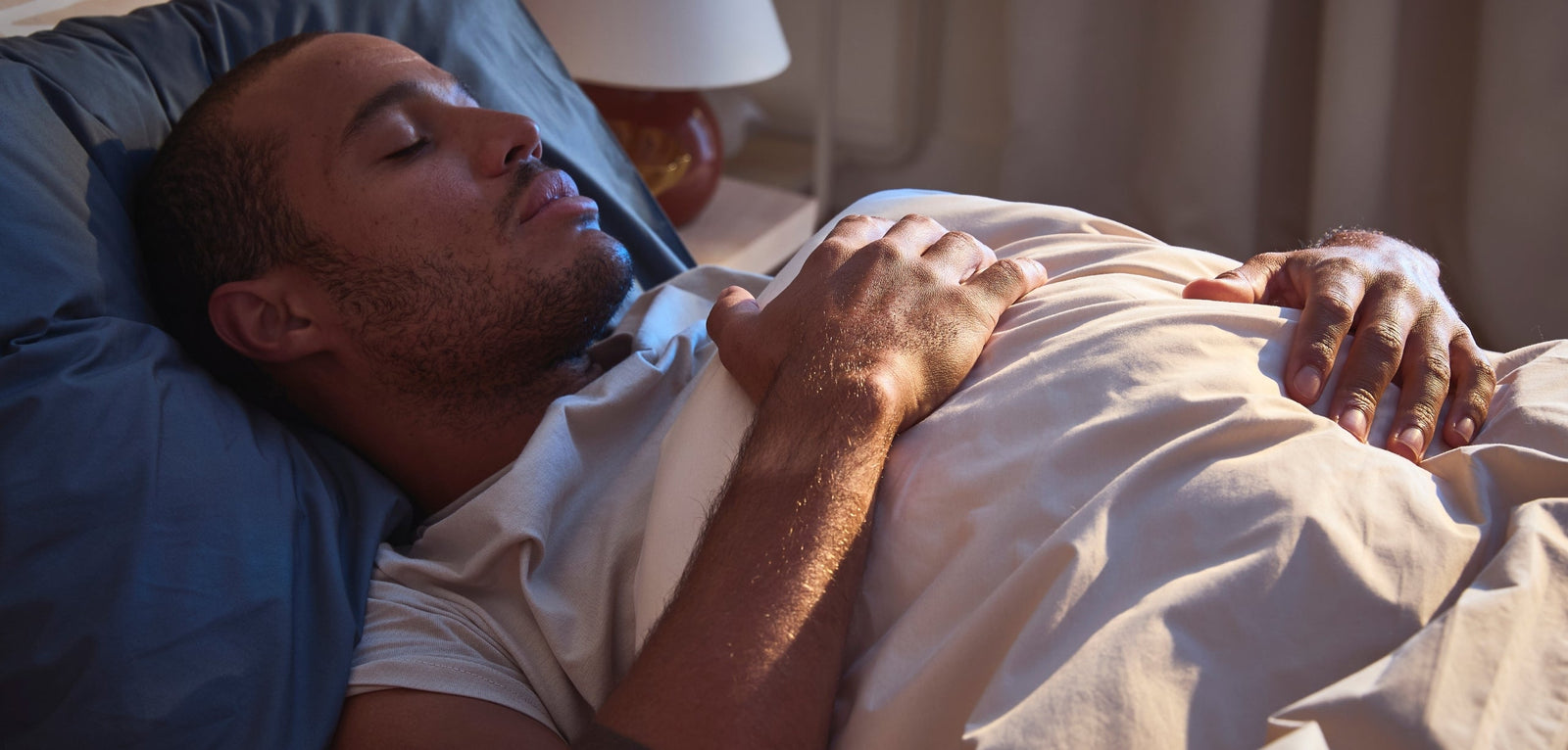 Un homme allongé sur le dos dans son lit, les yeux fermés et les mains posées sur la couette, dans une chambre éclairée par une lumière douce en fin de journée.