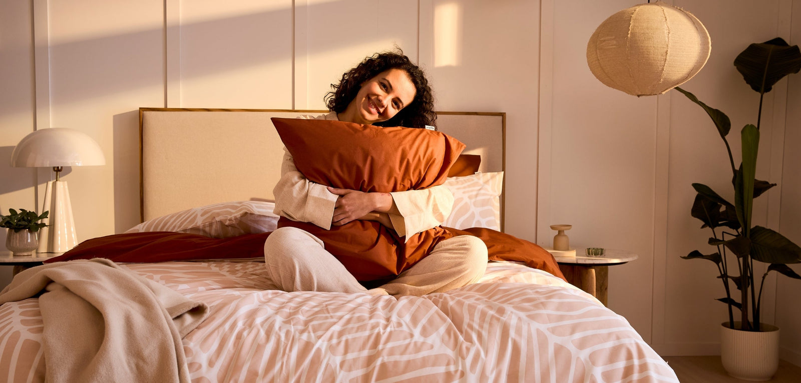 Une femme aux cheveux bouclés assise sur un lit, serrant un oreiller terracotta et souriant, dans une chambre décorée d'une plante verte et d'une suspension en osier.